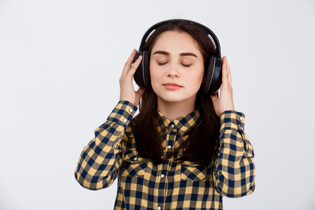 Young beautiful brunette girl dressed in plaid shirt listening music in headphones over white backgroundの写真素材