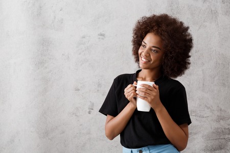 Young beautiful african girl holding cup, smiling over light background. Copy space.