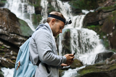 Young handsome man with backpack looking at phone, waterfall background.の写真素材