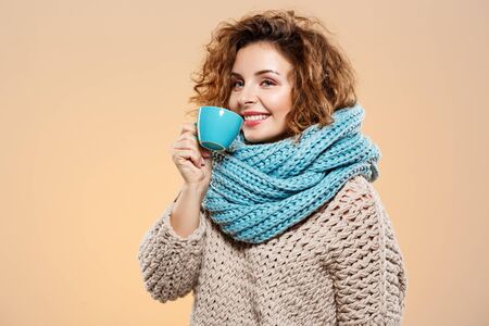 Close up portrait of cheerful smiling beautiful brunette curly girl in knitted sweater and grey neckwarmer holding cup over beige background.の写真素材
