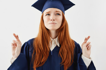 Portrait of beautiful yet nervous redhead graduate girl in mantle praying to success as future lawyer or doctor fingers crossed over white background.の写真素材