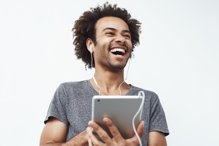 Portrait of cheerful happy african man in headphones laughing holding tablet talking or watching and enjoying a comedy show or browsing over white background.の写真素材