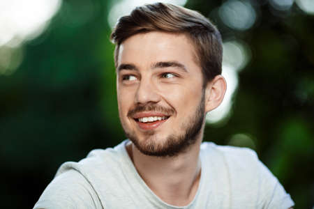 Close up portrait of handsome smiling young man in white t-shirt looking away on blurry outdoor...