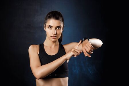 Young beautiful sportive girl pointing at watch over dark background.