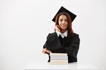 African female graduate in cap and mantle smiling sitting with books over white background.の写真素材