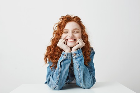 Pleased redhead girl smiling rejoicing with hands on cheeks sitting at table over white background.の写真素材