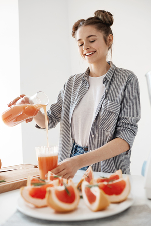 Happy beautiful girl pouring grapefruit detox smoothie in glass smiling over white wall. Healthy diet nutrition.の写真素材