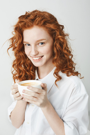 Portrait of tender redhead girl with freckles smiling holding cup looking at camera. White background.の写真素材