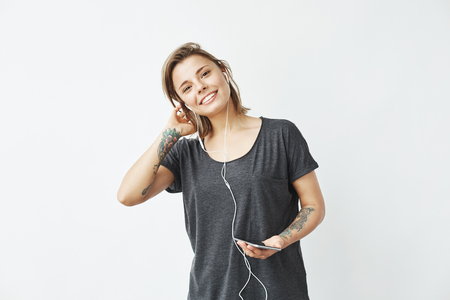 Happy young beautiful girl in headphones holding phone looking at camera smiling over white background.の写真素材