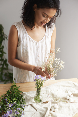 Young african female florist smiling working with bunch of flowers at workplace over white wall.の写真素材