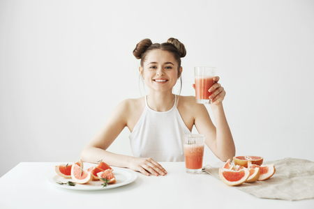 Happy beautiful girl smiling sitting at table holding glass with healthy detox fresh grapefruit smoothie over white wall.の写真素材