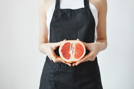 Girl in black apron holding half of grapefruit in hands over white wall. Copy space.の写真素材