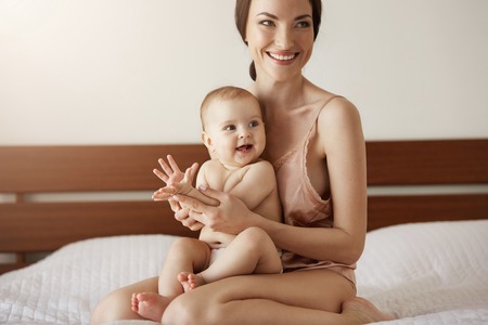 Young beautiful happy mother in sleepwear and her newborn baby sitting on bed in morning smiling playing together.の写真素材