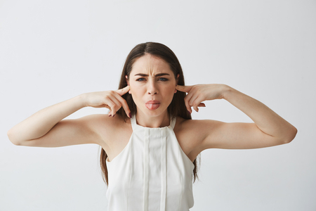 Displeased beautiful brunette girl closing ears with fingers from noise showing tongue over white background.の写真素材