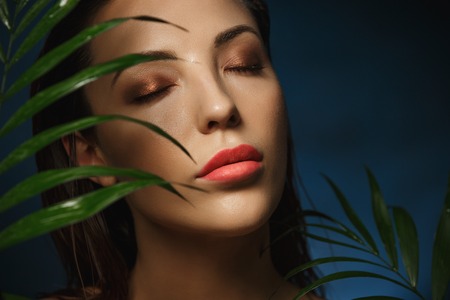 Beautiful woman face under exotic green leaves. Fashion photography.