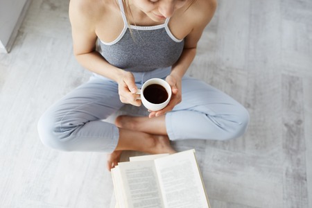 Portrait of tender young beautiful girl reading book holding cup of coffee sitting on floor over white wall. From above.の写真素材