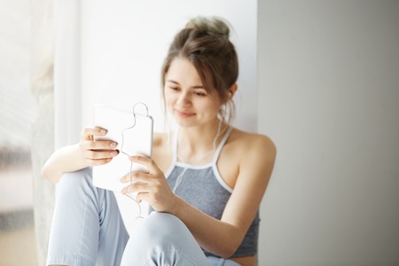 Portrait of young teenage cheerful girl in headphones smiling looking at tablet surfing web browsing internet sitting near window over white wall.の写真素材