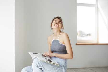 Portrait of young beautiful happy girl smiling laughing holding book sitting on chair over white wall at home.の写真素材