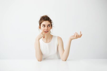 Dreamy beautiful young girl with bun sitting at table over white background thinking dreaming.