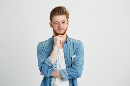 Portrait of confident young handsome man with beard looking at camera thinking with hand on chin over white background.の写真素材
