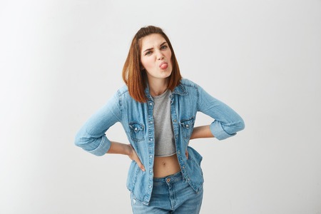 Young playful pretty girl looking at camera showing tongue with arms akimbo over white background.