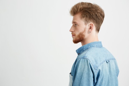 Portrait of young handsome man with beard wearing jean shirt standing in profile over white...