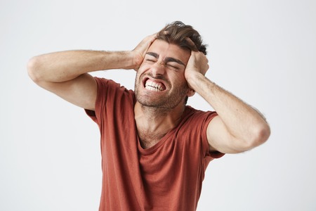 Emotional angry muscular man wearing red t-shirt closing eyes tight and screaming in pain or full...