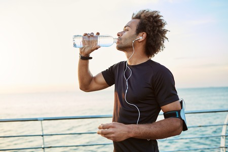 Slim and young sportsman refreshing himself, drinking water out of plastic bottle in the morning sunlight. Listening to his favourite songs while jogging. Young Afro-American practicing healthy lifestyleの写真素材