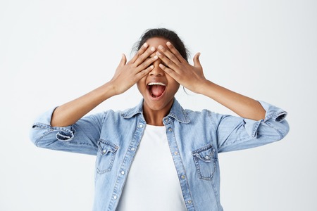 Positive cheerful Afro-American female dressed casually holding her hands on closed eyes with open in excitement mouth expecting for surprise or gift. People, good news, positive emotions.の写真素材