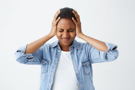Photo of disappointed Afro-american woman in denim shirt holding her hands on temples frowning face and closed eyes in despair and terror. Woman regreting her act. Female in despair and shockの写真素材