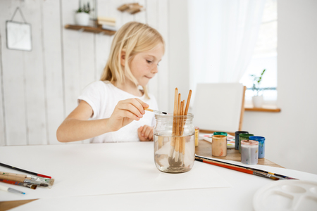 ?reative little blonde girl with freckles washing brush in jar of water during art lesson. Cute child concentrated on painting. Kids art and activities.の写真素材