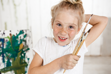 Portrait of angel-like cheerful smiling with teeth child in white morning light in art room, holding in her hand bunch of brushes. Little European girl with blond hair looking happy and joyful showing positive emotions.の写真素材