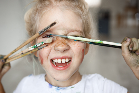 Blonde little girl in playful mood with paint on her freckled face and blue eyes covering her face with brushes and looking through them at you like hiding. Playing, smiling child showing her teeth. Positive emotions.の写真素材