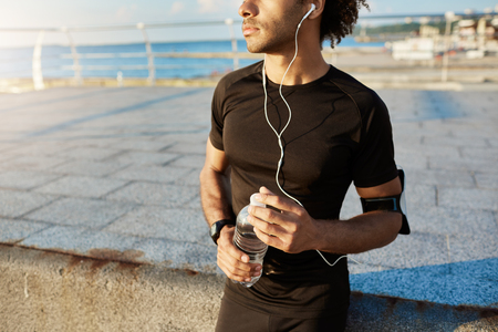 Midsection of male dark-skinned runner in black sportswear holding bottle of mineral water in his hands, using music app on mobile phone during jogging workout behind the sea.の写真素材