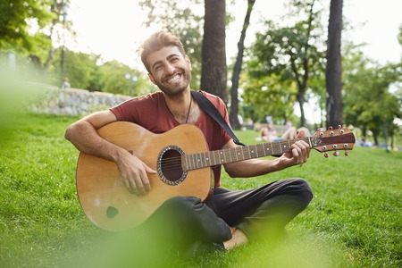 Positive bearded guy with dark warm eyes dressed casually having pleasant smile showing his perfect white teeth playing guitar, sitting cross-legged on grass, having good mood.の写真素材