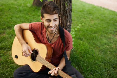 Talented attractive male musician playing guitar in the park under the tree, having good time, sitting at grass enjoying splendid weather. Romantic young man love singing songs.の写真素材