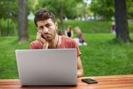Upset man with beard and stylish hairstyle wearing casual red T-shirt holding his hand on chin having tired and unhappy look. Student sitting at the desk with notebook and smartphone, tired of studying. People, emotions, body language and lifestyle concepの写真素材