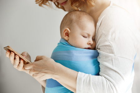 Beautiful young mother reads articles about maternity life on phone while little lovely son sleeping on her chest in blue baby sling.の写真素材