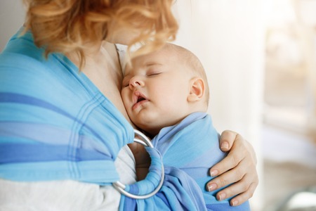 Close up portrait of innocent newborn boy having sweet dreams on mother chest in baby sling. Mom looking at her child with love and tenderness.の写真素材