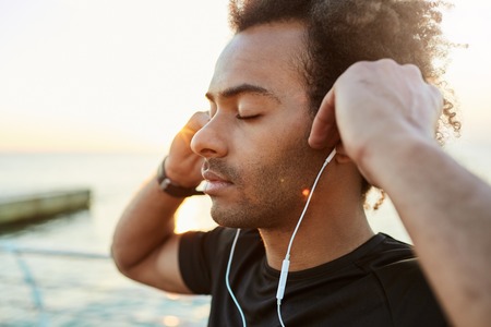 Portrait of mediative and peaceful Afro-American runner with bushy hairstyle and closed eyes listening music. Outdoor shot of dark-skinned sportsman in black T-shirt relaxing after morning workout session by the sea.の写真素材
