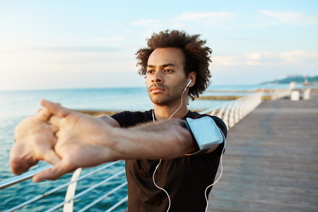 Afro-American male runner with beautiful athletic body and bushy hair stretching muscles, raising his arms while warming up before morning workout session. Young man athlete exercising outdoors.の写真素材