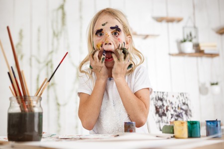 Playful Caucasian female child having fun with spots of paint on her face and opened mouth, touching her face with hands in paint. Little messed up in paint blonde girl having fun and playing fool.の写真素材