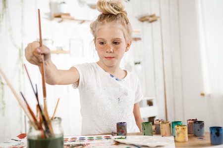 Indoor shot of European cute creative child with hair bun snd blue eyes occupied with drawing. Little blonde girl wearing white t-shirt busy with painting, deeping brush into water, drawing new masterpiece for her parents.の写真素材
