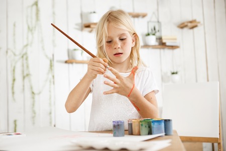Cute, beautiful blonde girl in white t-shirt joyfully painting her palm with brush, standing behind the desk with jar of water, brushes, and paint on it. Isolated shot, horizontal.の写真素材