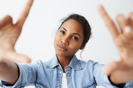 Close up studio shot of beautiful charming brunetteAfro-American model with healthy dark skin looking at camera with faint smile, posing indoors against blank wall background, wearing denim shirt.の写真素材