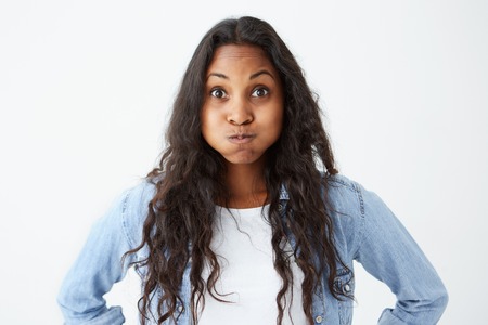 Charming mocking Afro-american woman wearing loose wavy hair looking with bugged eyes at the camera, with full mouth. Dark-skinned model posing on isolated white background.の写真素材