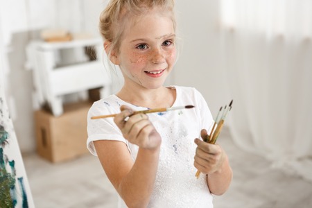Close-up portrait of blonde cute European little girl with paint on her freckled face and hair bun smiling with all her teeth holding a bunch of brushes in her hands. Cheerful girl messed up her white t-shirt, enjoying art activities.の写真素材