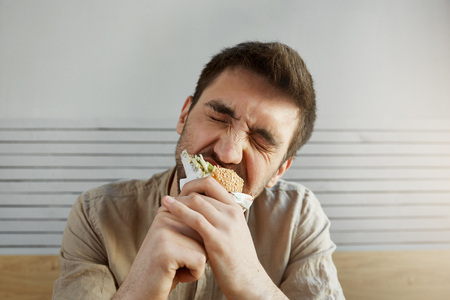 Young unshaven handsome guy with dark hair eating sandwich in fast food with closed eyes, with happy and satisfied expressionの写真素材