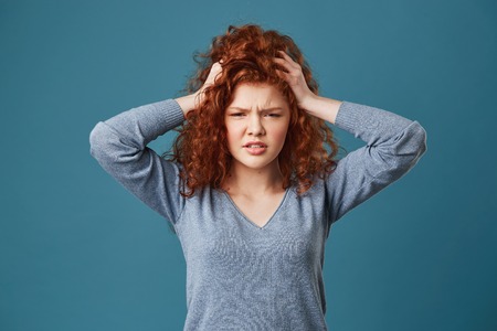 Portrait of young ginger girls with wavy hair and freckles holding hands on head being tired of head...