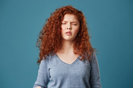 Close up of pretty young girl with wavy ginger hair and freckles in grey shirt having headache after sleepless nightの写真素材